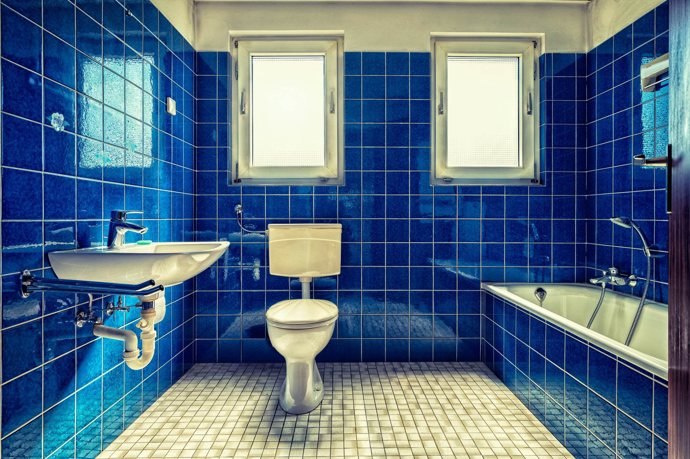 Salle de bain ancienne entièrement carrelée de bleu vif, lavabo, baignoire et deux petites fenêtres