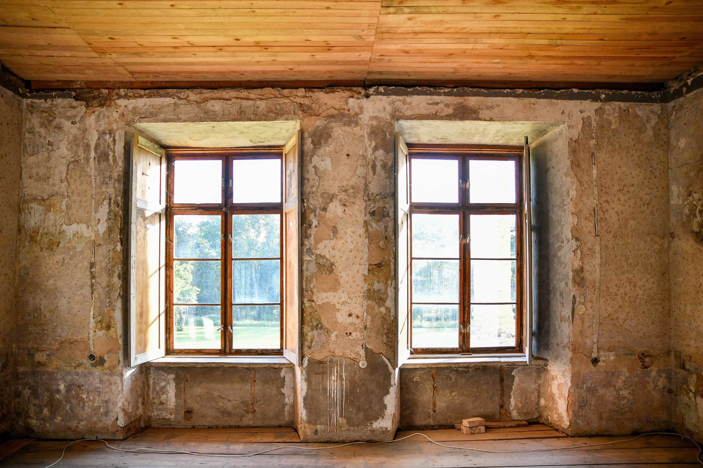 Pièce vidée avant travaux, murs décroûtés jusqu’à la maçonnerie, plafond en bois et deux fenêtres anciennes