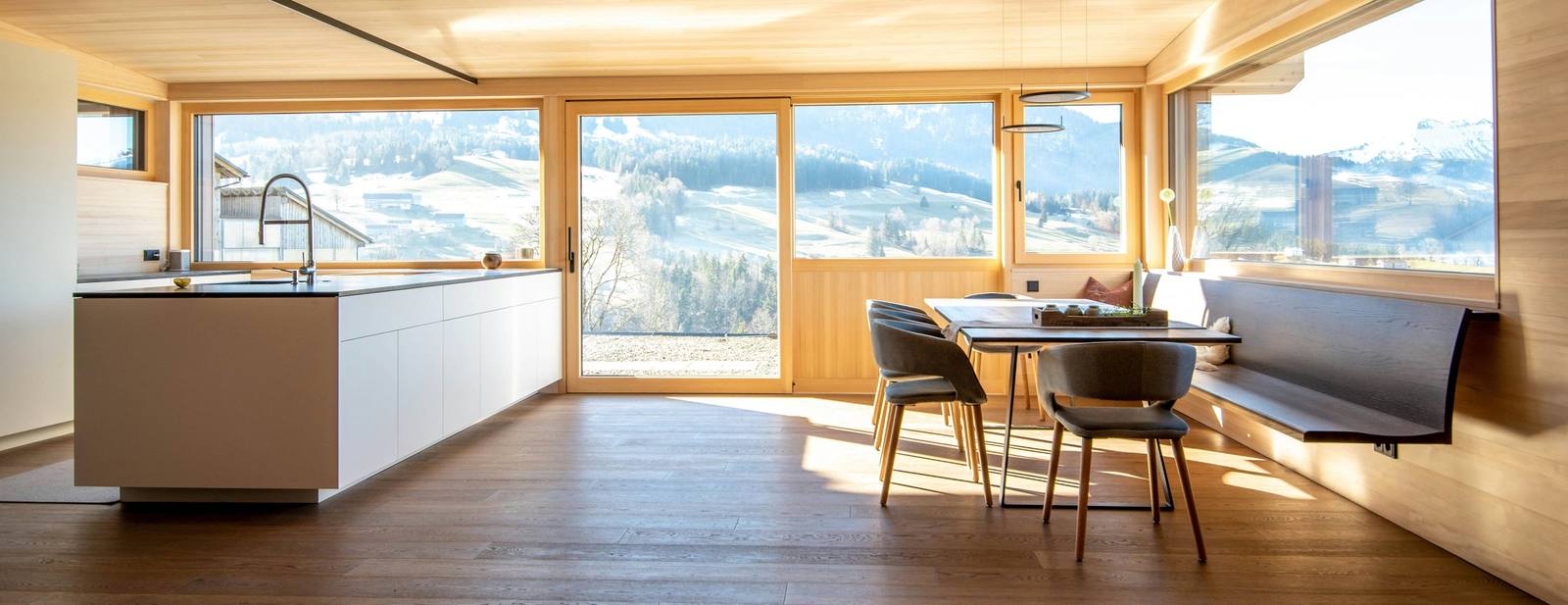 Séjour d’un chalet rénové, bois clair au plafond, coin repas et grandes baies ouvertes sur les pâturages enneigés et la montagne